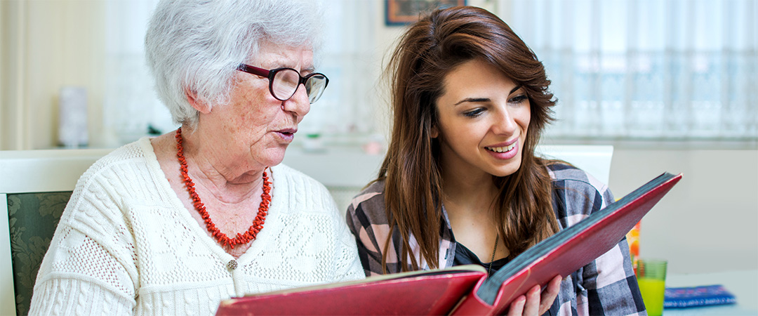 Senior woman with red necklace looking through a photo scrapbook with a younger woman with long brown hair.